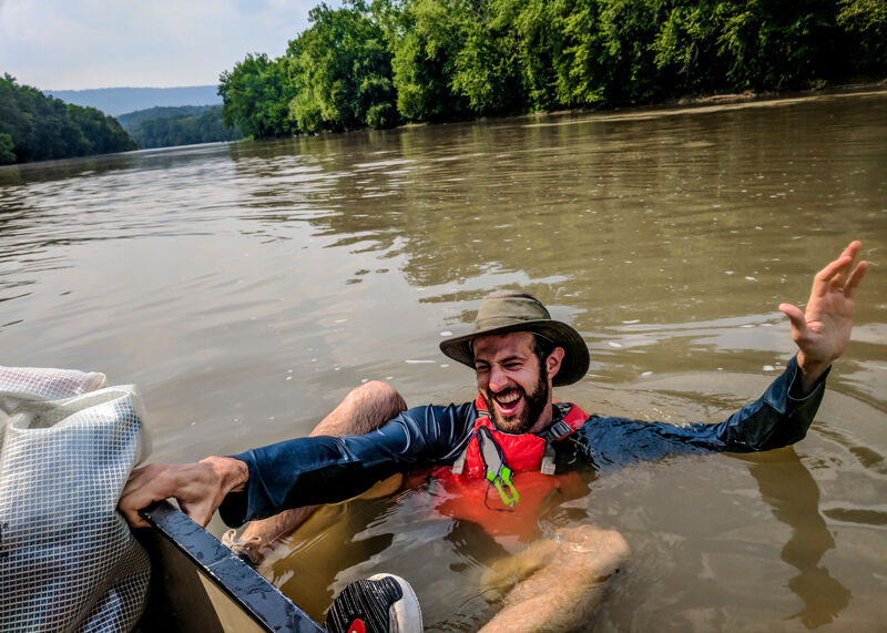 A man is in the water next to a canoe. He is wearing a hat and a life vest. He is smiling and waving. The canoe is partially submerged. The water is murky and there are trees in the background. The man seems to be enjoying himself despite the situation.